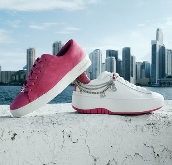 Dos zapatillas sobre una repisa junto al agua: una zapatilla de ante rosa y una zapatilla blanca con plataforma, con cadenas plateadas y adornos de corazones. Fondo con el skyline urbano.