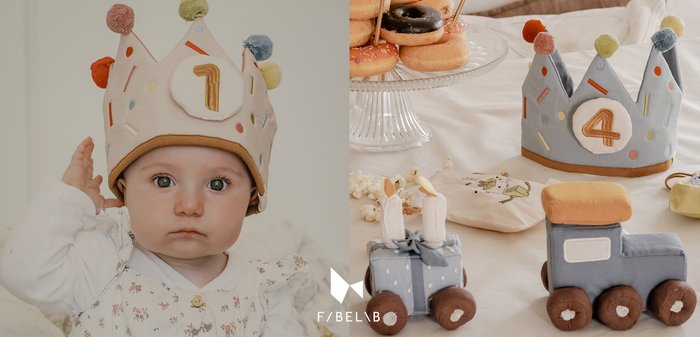 Két szövetkorona színes pompomokkal, egy bézs az "1" számmal és egy kék a "4" számmal, plüss játékokkal és fánkokkal egy elegáns asztalon.