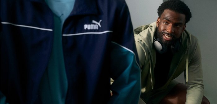 Mann mit Kopfhörern um den Hals beugt sich vor und spricht mit einer anderen Person, die eine blaue Puma-Jacke trägt, in einem Innenraum.