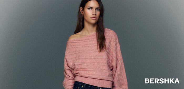 Růžový pletený svetr s texturovanými horizontálními vzory, s vykrojením odhalujícím ramena a žebrovaným lemem. Kombinován s tmavými džínami.