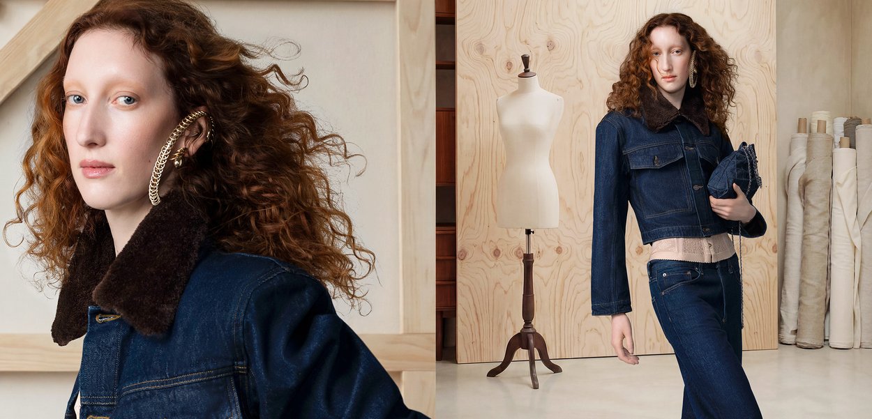 Mujer con cabello rizado rojo lleva chaqueta vaquera oscura y vaqueros, sosteniendo denim doblado en estudio con maniquí y rollos de tela al fondo.