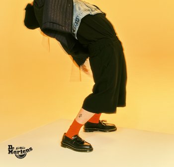 Zapatos de charol negros con punta redonda, combinados con calcetas de color naranja brillante. Unos pantalones cortos negros holgados y una chaqueta de mezclilla complementan el conjunto.
