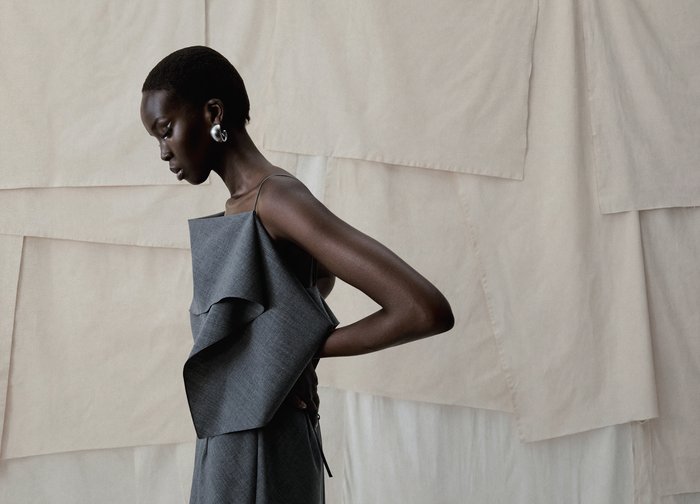 Γυναίκα με κοντά μαλλιά φοράει ασύμμετρο γκρι φόρεμα και μεγάλα ασημένια σκουλαρίκια, ποζάροντας πλαγίως μπροστά σε μπεζ υφασμάτινο φόντο.