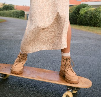 Jupe beige à motifs, bottes à lacets beige clair avec une semelle texturée, se tenant sur un skate en bois avec des roues sur une surface pavée.