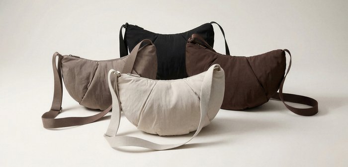 Vier halbmondförmige Stoff-Schultertaschen in Schwarz, Braun, Taupe und Creme auf neutralem Hintergrund angeordnet.