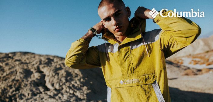Young person adjusts hood on yellow Columbia windbreaker outdoors in sunny, rocky landscape with clear blue sky.