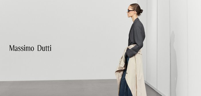 Donna con occhiali da sole, blazer scuro, trench beige legato in vita e jeans a gamba larga, in piedi in una stanza minimalista con il testo "Massimo Dutti".