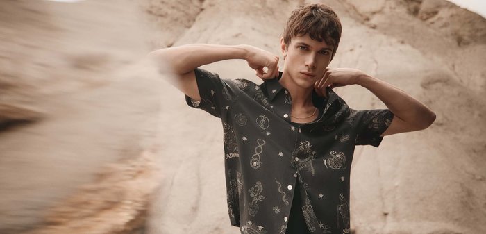 Junger Mann richtet den Kragen eines schwarzen Kurzarmhemds mit himmlischen und tierischen Mustern, stehend in einer felsigen Außenumgebung.