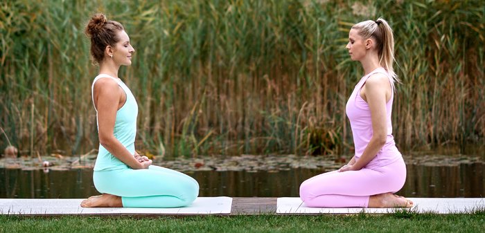 Twee vrouwen knielen op yogamatten bij een vijver, gekleed in pastelgroene en roze tanktops en leggings. Op de achtergrond zijn hoge grassen en water te zien.