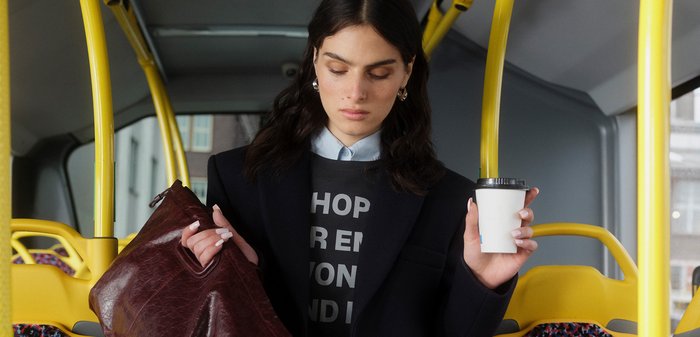 Sac en cuir marron foncé, sweat gris avec texte, blazer noir, chemise bleu clair, tenant une tasse de café blanche, intérieur de bus jaune.