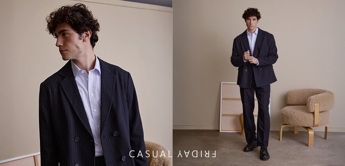 Dunkler Nadelstreifenanzug mit zweireihigem Sakko und gerade geschnittenen Hosen, kombiniert mit einem weißen Hemd. Schwarze Schuhe und ein strukturiert-cremiger Sessel im Hintergrund.