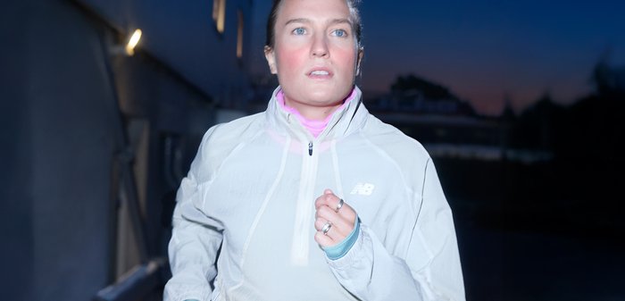 Femme courant à l'extérieur au crépuscule, portant une veste New Balance de couleur claire et affichant une expression concentrée.