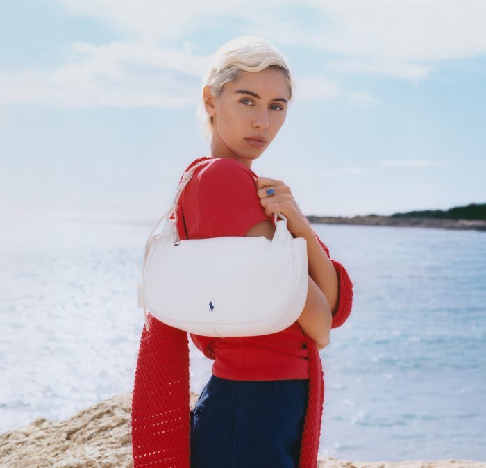 Young woman with short blonde hair, wearing red top and navy pants, holding white handbag by the sea on a sunny day.