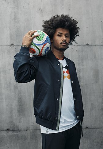 Czarna kurtka bomberka z srebrnymi akcentami, noszona na białej koszulce z pomarańczowo-czarnymi wzorami w romby, gracz trzymający kolorową piłkę nożną.