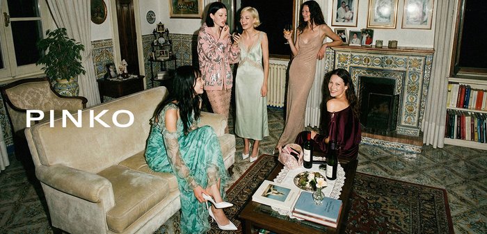 Pięć kobiet ubranych w eleganckie suknie wieczorowe towarzyszy sobie w przytulnym salonie, przy drewnianym stoliku kawowym, na którym znajdują się wino i książki.