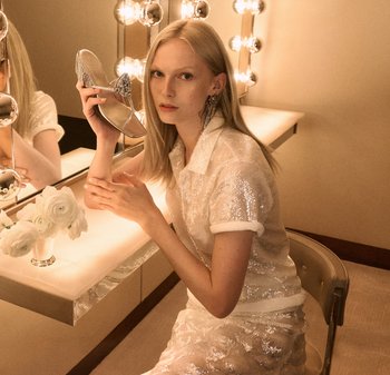 Silverfärgad högklackad sandal med strassdekorationer. Modell i en paljettklädd vit topp håller skon och sitter vid en upplyst sminkbord.