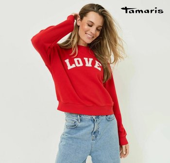 Woman with long hair smiling, wearing a red sweater with "LOVE" text and light blue jeans, standing against a plain white background.