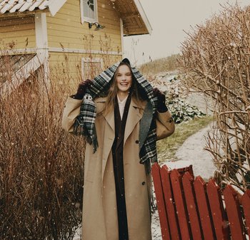 Beige trenchcoat, grijze en geruite sjaal, bordeauxrode handschoenen. Sneeuwrijke buitenomgeving met houten hek en struiken op de achtergrond.