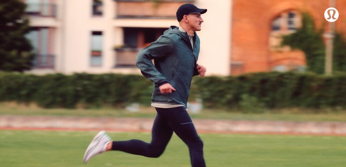 Groene lichtgewicht hardloopjack over een licht shirt, zwarte leggings en witte sneakers. De achtergrond toont woongebouwen en groen.