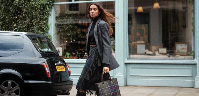 Veste gris par-dessus un col roulé noir, jupe à carreaux avec franges, portant un sac à main à carreaux. Vue de côté d'une voiture noire dans un cadre urbain.