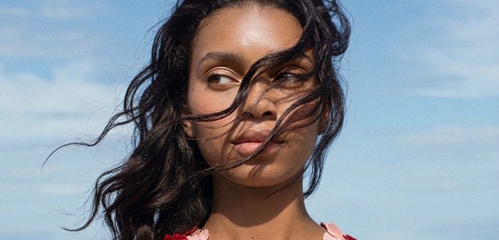 Femme aux longs cheveux foncés flottant sur son visage, regardant sur le côté, avec un ciel bleu clair en arrière-plan.