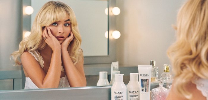 Produkte für die Haarpflege von Redken sind ausgestellt: zwei weiße Flaschen und eine weiße Tube mit bunten Etiketten. Das Model hat langes, welliges blondes Haar und helle Haut.