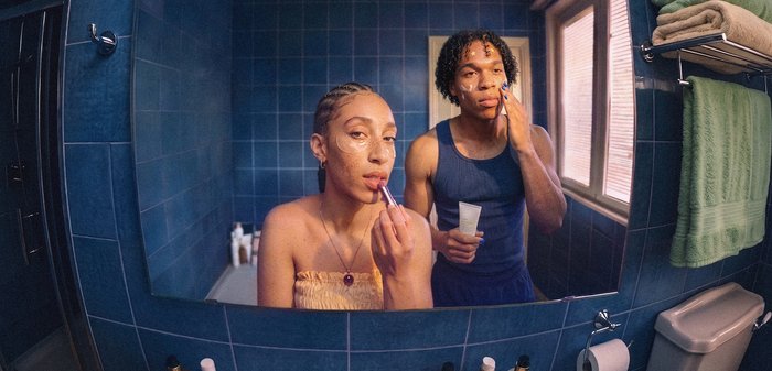 Deux personnes appliquent ensemble des soins de la peau et du maquillage dans une salle de bain carrelée de bleu, utilisant un miroir au-dessus de l'évier avec des serviettes sur un porte-serviettes à proximité.