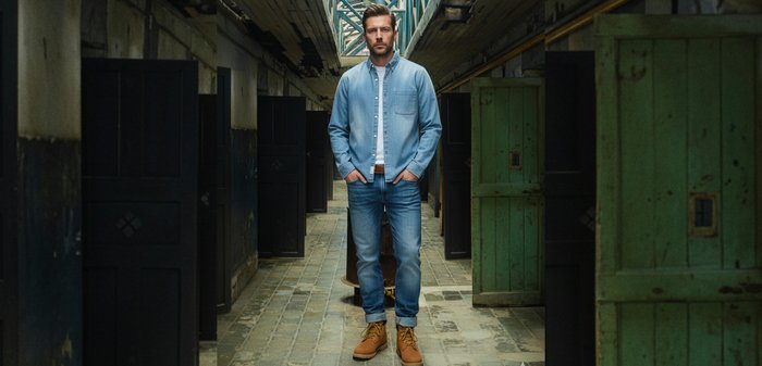 Lichtblauw denim overhemd en bijpassende spijkerbroek, bruine riem en beige laarzen. De omgeving heeft versleten muren en groene houten deuren.