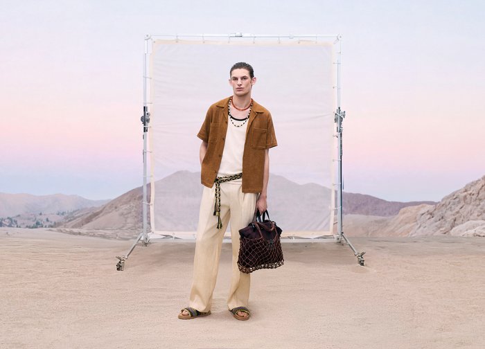 Jonge man in beige broek en bruin overhemd die een grote tas vasthoudt, staand in een woestijnachtig landschap met een stoffen achtergrond achter hem.