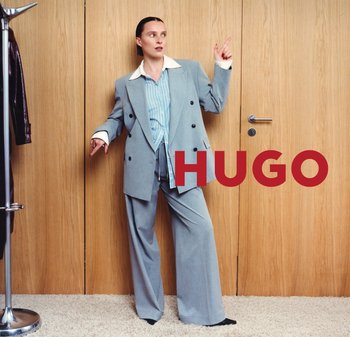 Mujer con traje gris oversize y camisa de rayas posa con los dedos señalando hacia arriba y hacia abajo frente a una pared de madera con el texto "HUGO".
