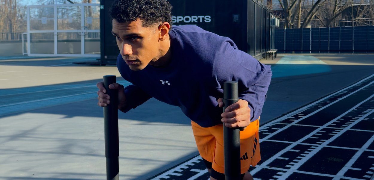 Man in purple shirt and orange shorts gripping black bars, leaning forward, preparing for a workout on an outdoor sports court.