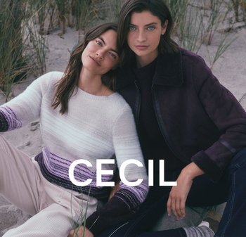 Zwei Frauen sitzen auf sandigem Boden. Eine trägt einen weißen und lila Pullover mit gestreiften Mustern, die andere eine dunkle Jacke mit einem Fellkragen.