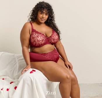 Krøllet-haired kvinde sidder på hvidt opdækket underlag med røde rosenblade, iført rød blonde-bh og matchende højtaljede trusser.