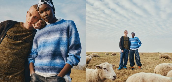 Deux mannequins se tiennent dans un champ avec des moutons, portant un pull rayé gris et bleu et un pull à motifs vert foncé, avec des chaussures bleues et orange.