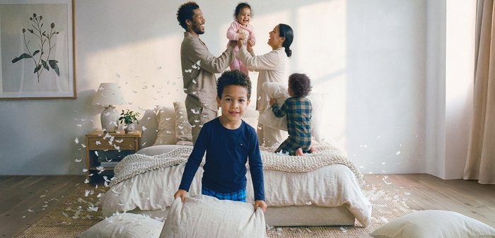 Μια ζεστή σκηνή υπνοδωματίου με ανοιχτόχρωμο, υφασμάτινο κάλυμμα κρεβατιού και πολλά διακοσμητικά μαξιλάρια. Ένα ξύλινο κομοδίνο φιλοξενεί μια γλάστρα με φυτό.