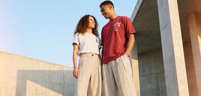 Deux personnes se tiennent debout en se souriant, portant des pantalons et t-shirts décontractés aux tons neutres à côté d’une structure moderne en béton sous un ciel dégagé.