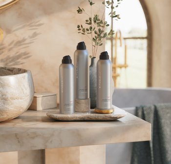 Trois canettes de mousse de douche en métal argenté avec des capuchons noirs sur un plateau en marbre beige. Une serviette verte texturée et un bol rond en pierre sont à proximité.
