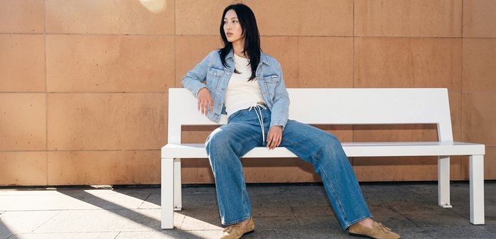 Jeune femme portant une veste en jean et un jean, assise détendue sur un banc blanc devant un mur carrelé beige à l'extérieur.