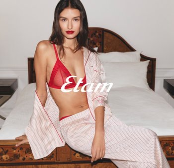 Frau in roter Unterwäsche und weißer gepunkteter Pyjamahose sitzt auf einem Holzbett mit weißer Bettwäsche in einem minimalistischen Schlafzimmer.