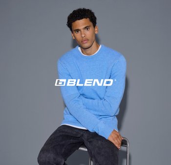 Pullover azzurro chiaro in maglia con scollatura rotonda, indossato sopra una camicia bianca, abbinato a jeans scuri. Modella seduta su uno sgabello, sfondo neutro.