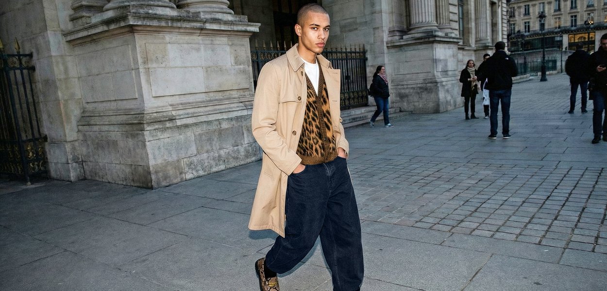 Mann går på bygate iført beige trenchcoat, leopardmønstret genser, mørke bukser og mønstrede sko nær et historisk bygg.