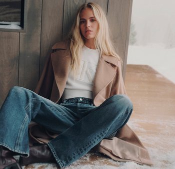 Ung kvinna med långt blont hår som bär en beige kappa, vit tröja, blå jeans och mörka stövlar, sittande inomhus vid ett fönster med snö utanför.