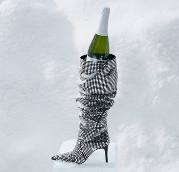 Svart høye hæler støvlett dekket av rhinestones, med et rynket design, holder en champagneflaske, satt mot en snødekt bakgrunn.