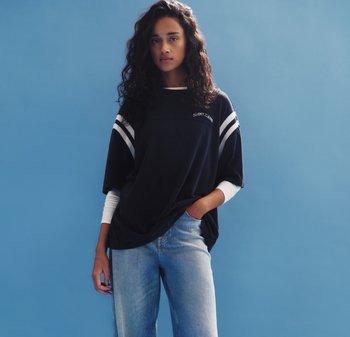 Jonge vrouw met krullend haar, gekleed in een oversized zwart Tommy Jeans-shirt over een wit shirt met lange mouwen en lichtblauwe spijkerbroek, staand tegen een blauwe muur.