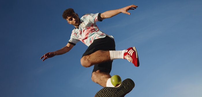 Jeune homme en l'air équilibrant une pomme verte sur la semelle de sa chaussure sur fond de ciel bleu clair.