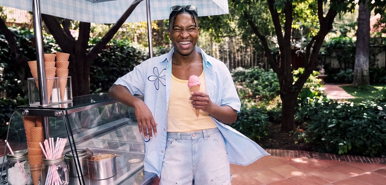 Persona sonriente está junto a un carrito de helados al aire libre sosteniendo un cucurucho de helado rosa, lleva pantalones cortos, una camisa azul claro y gafas de sol en la cabeza.