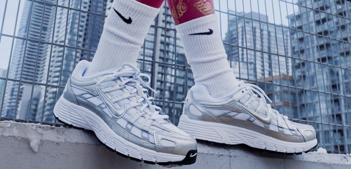 Vita och silverfärgade Nike-sneakers med vita Nike-strumpor, båda bärna av en person som sitter på en betongkant med en stadssilhuett i bakgrunden.