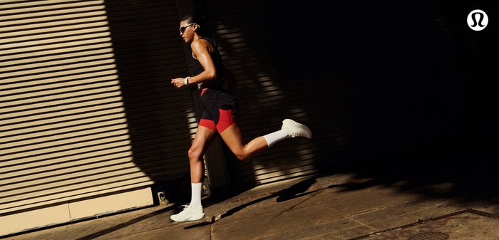 Runner wearing sunglasses, black and red athletic wear, and white shoes mid-stride on a sunlit urban street against a shuttered door.