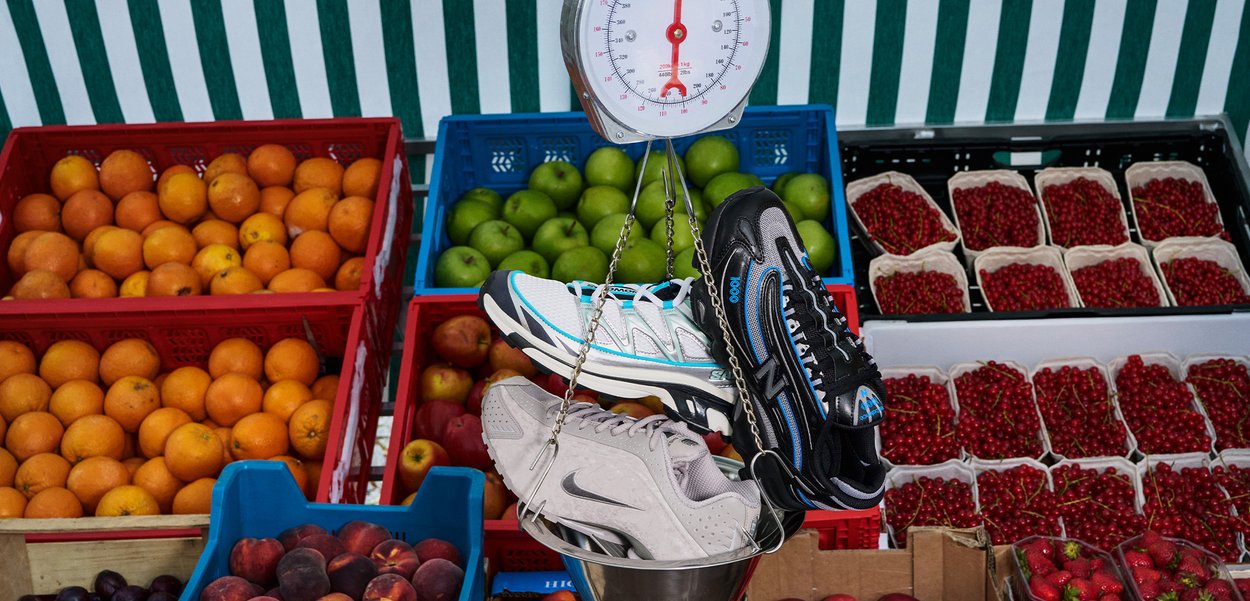 Deux chaussures de sport - l'une noire avec des accents bleus, l'autre gris clair - suspendues à une balance au-dessus de crates de fruits colorés dans un marché.