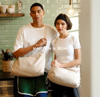 Mladý muž a žena v bílých topách a s béžovými křížovými taškami, stojící v kuchyni se zeleně obloženou zdí a visící žárovkou.